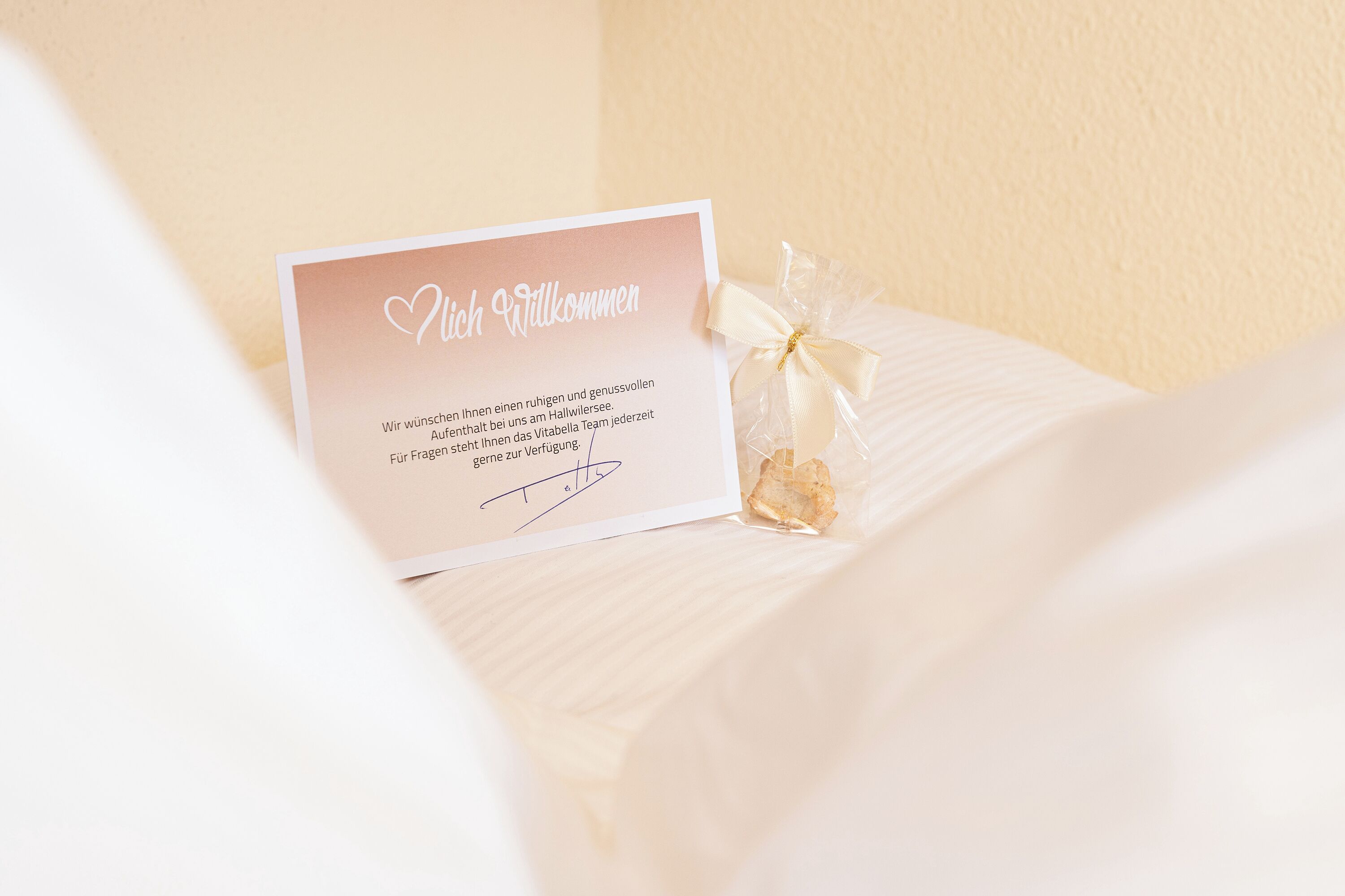 A welcome card with a heart symbol and a signature, placed on a bed with a white cover.