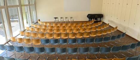 Un salón de conferencias con filas de sillas, una pantalla para proyector y un piano.