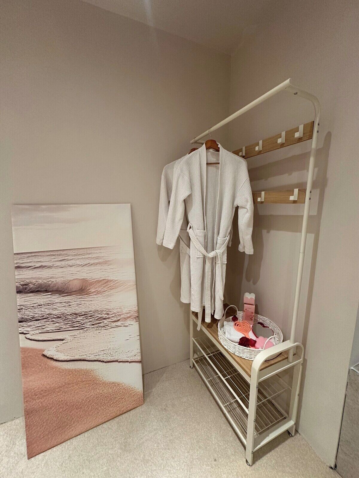 Une chambre avec un peignoir blanc sur un porte-manteau, un panier blanc contenant des articles de toilette et un tableau sur le thème de la plage.