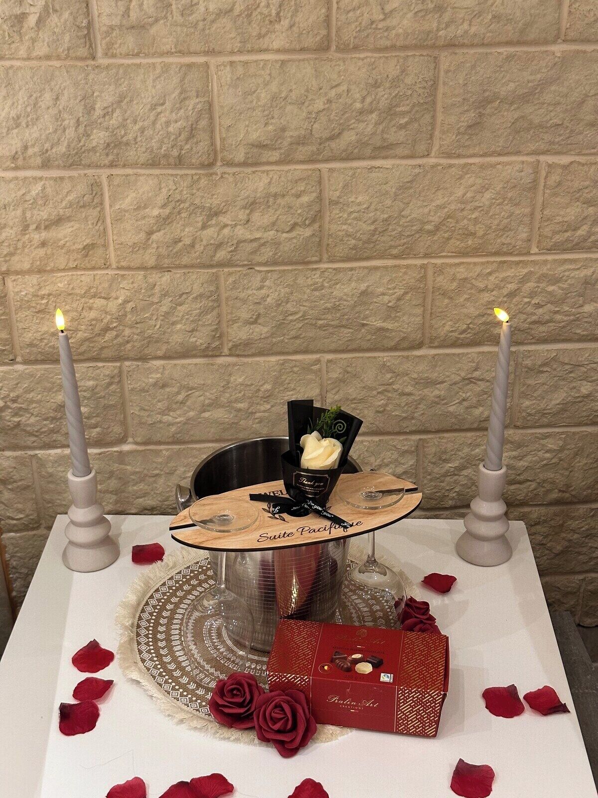 Ein Tisch mit Kerzen, einer Dankeskarte, einem Weinglas, Rosenblüten und einer Schachtel Pralinen.