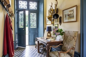 A room with a checkered floor, a blue door with decorative glass panels, a wooden table with a lamp, a chair, and wall-mounted decorations.