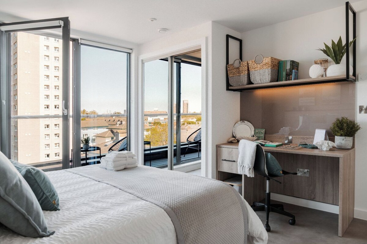 A modern bedroom with a large bed, a desk, and a balcony with a view of the city.