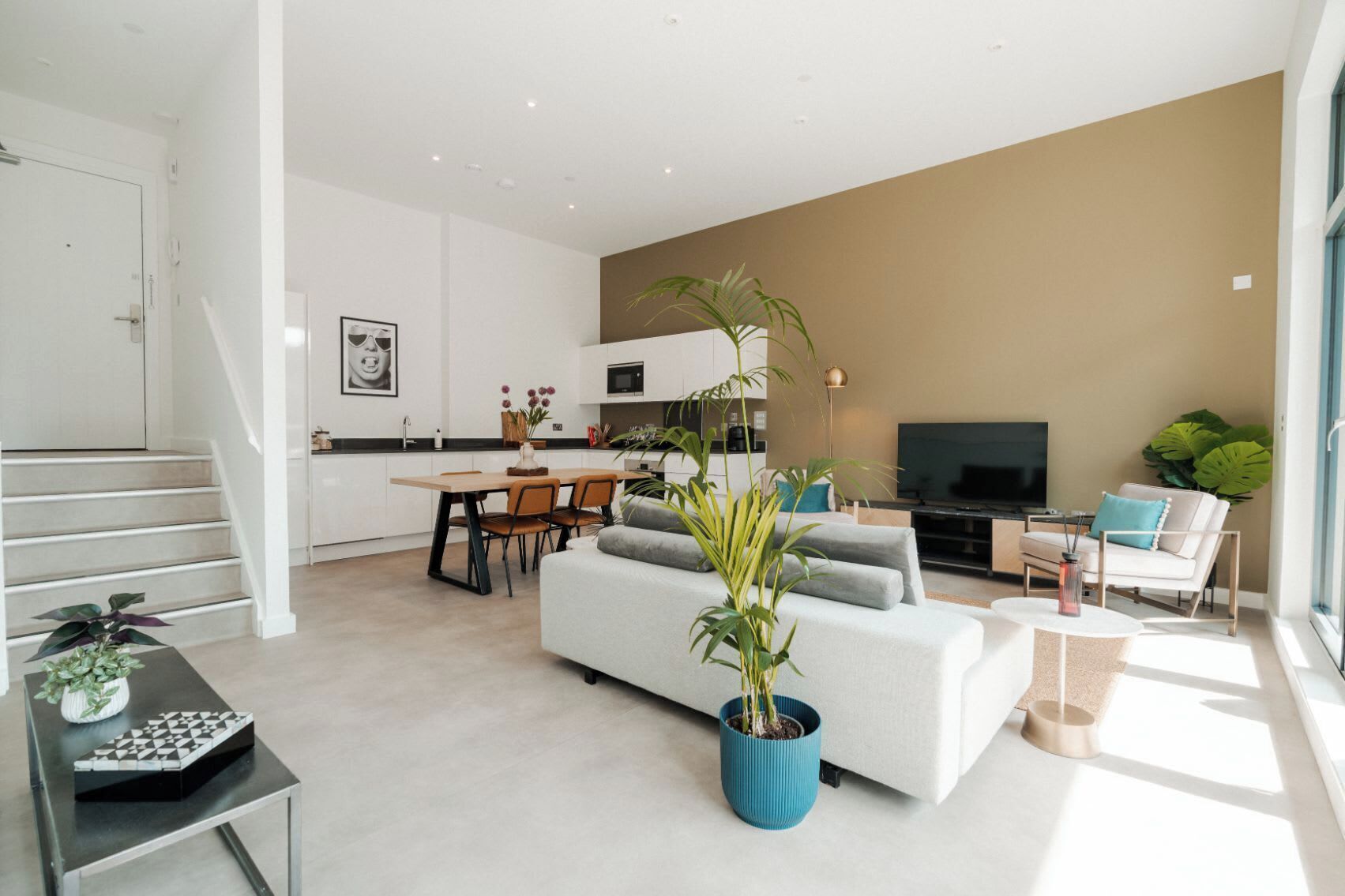 A modern living room with a sofa, a coffee table, and a TV. There is a staircase leading to a kitchen area with a dining table and chairs.