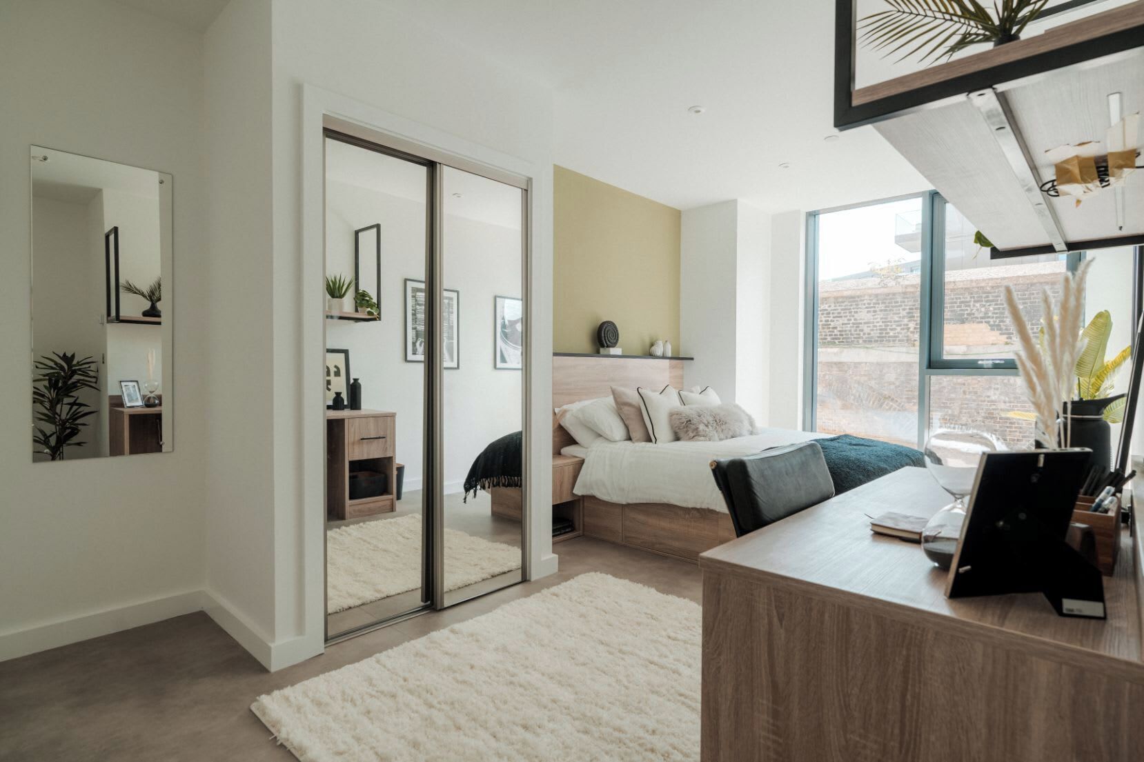 A modern bedroom with a bed, bedside table, and a desk with a computer. There is a sliding door leading to another room, and a large window providing natural light.
