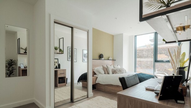 Een moderne slaapkamer met een bed, nachtkastje en bureau met computer. Er is een schuifdeur naar een andere kamer, en een groot raam zorgt voor natuurlijk licht.