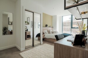 Ein modernes Schlafzimmer mit Bett, Nachttisch und Schreibtisch inklusive Computer. Eine Schiebetür führt in einen anderen Raum, und ein großes Fenster lässt viel natürliches Licht herein.