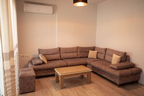 A living room with a sectional sofa, a wooden coffee table, and a ceiling light fixture. - Perla Apartment 02 in Plazhi San Pietro (Plazhi San Pietro)