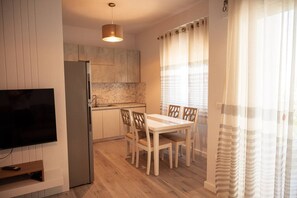A compact kitchen with a built-in refrigerator, a dining table with chairs, a TV mounted on the wall, and a window with curtains. - Perla Apartment 02 in Plazhi San Pietro (Plazhi San Pietro)