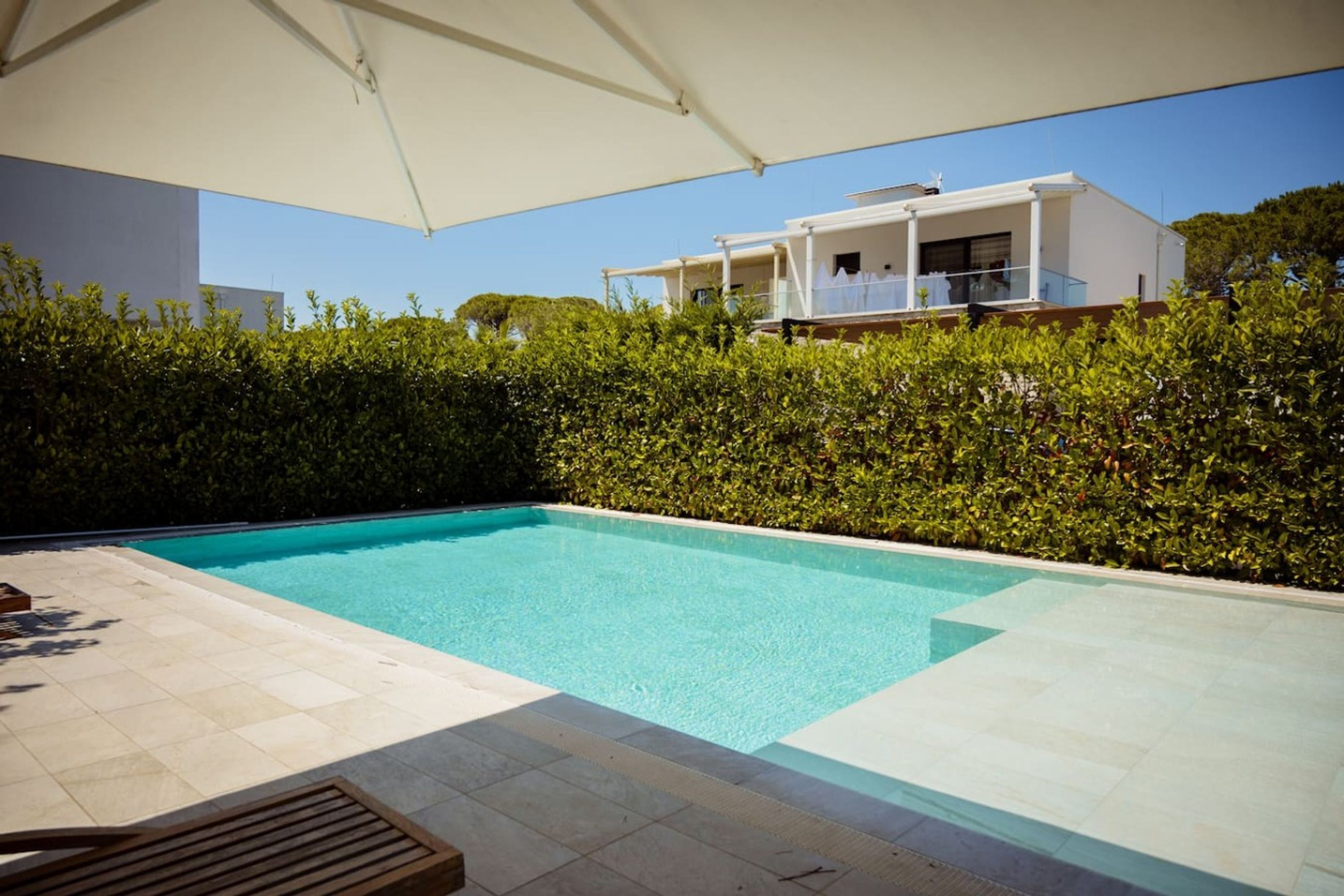 Une piscine dotée d’une terrasse carrelée, entourée d’une haie et bordée d’un bâtiment blanc avec un balcon.