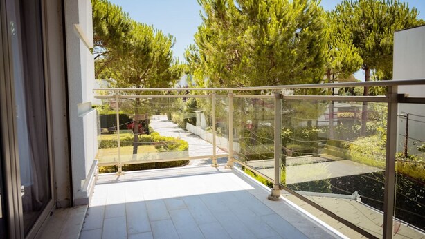 Een balkon met een glazen hekwerk, betegelde vloer en uitzicht op bomen en een tuin.