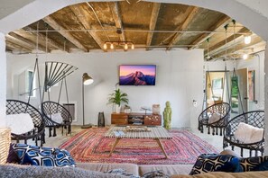 A spacious living area with a large rug, hanging chairs, a wooden cabinet, and a wall-mounted TV. - Eclectic Asheville Escape w Fire Pit & Yard! (Asheville)