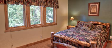 A bedroom with a wooden bed frame, a floral bedspread, a window with a valance, a nightstand with a lamp, and a framed picture on the wall.