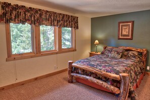A bedroom with a wooden bed frame, a floral bedspread, a window with a valance, a nightstand with a lamp, and a framed picture on the wall. - Moose Meadow Retreat (Blue River)