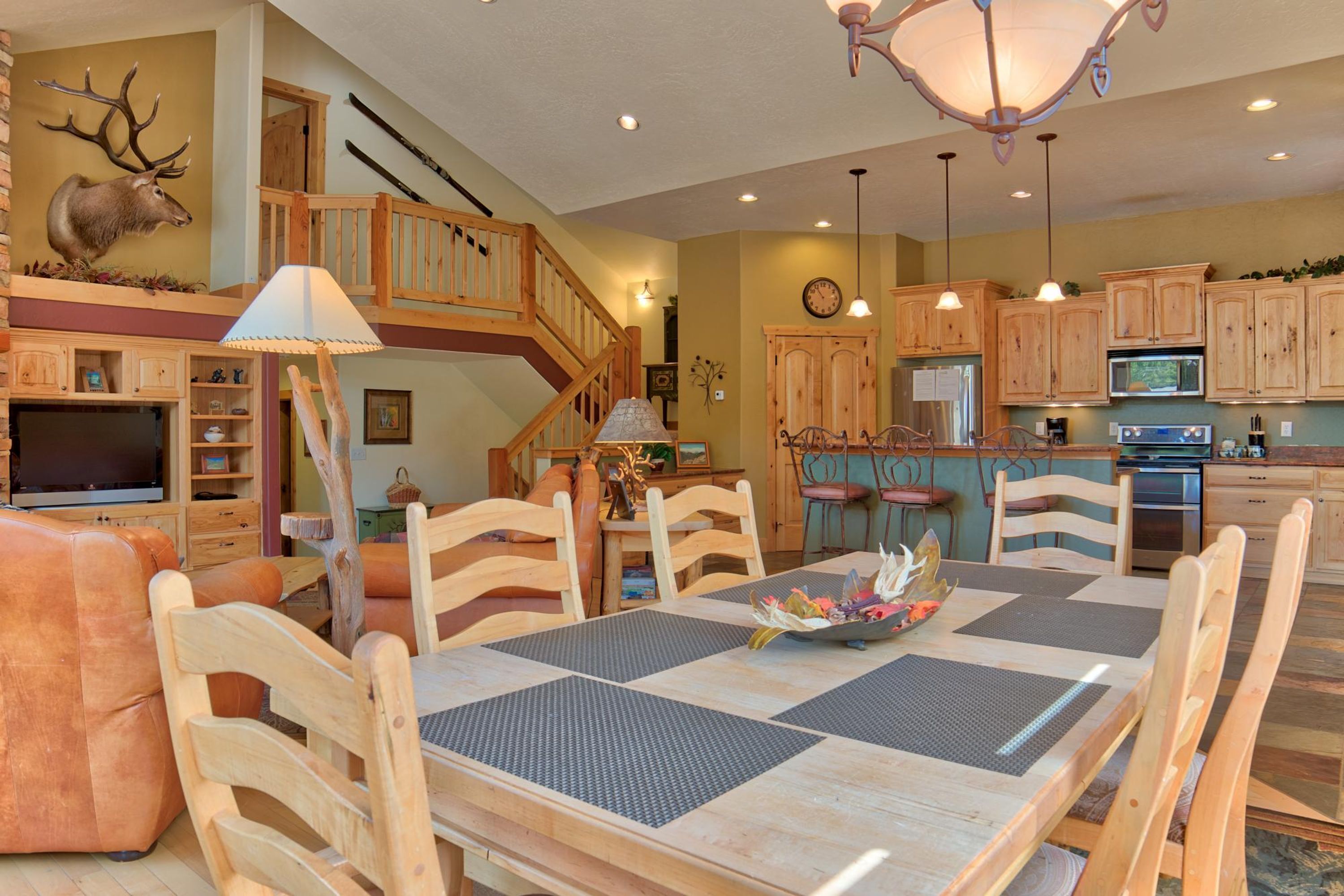 A spacious dining area with a high wooden table and chairs, a chandelier, and a kitchen with wooden cabinets and a refrigerator.