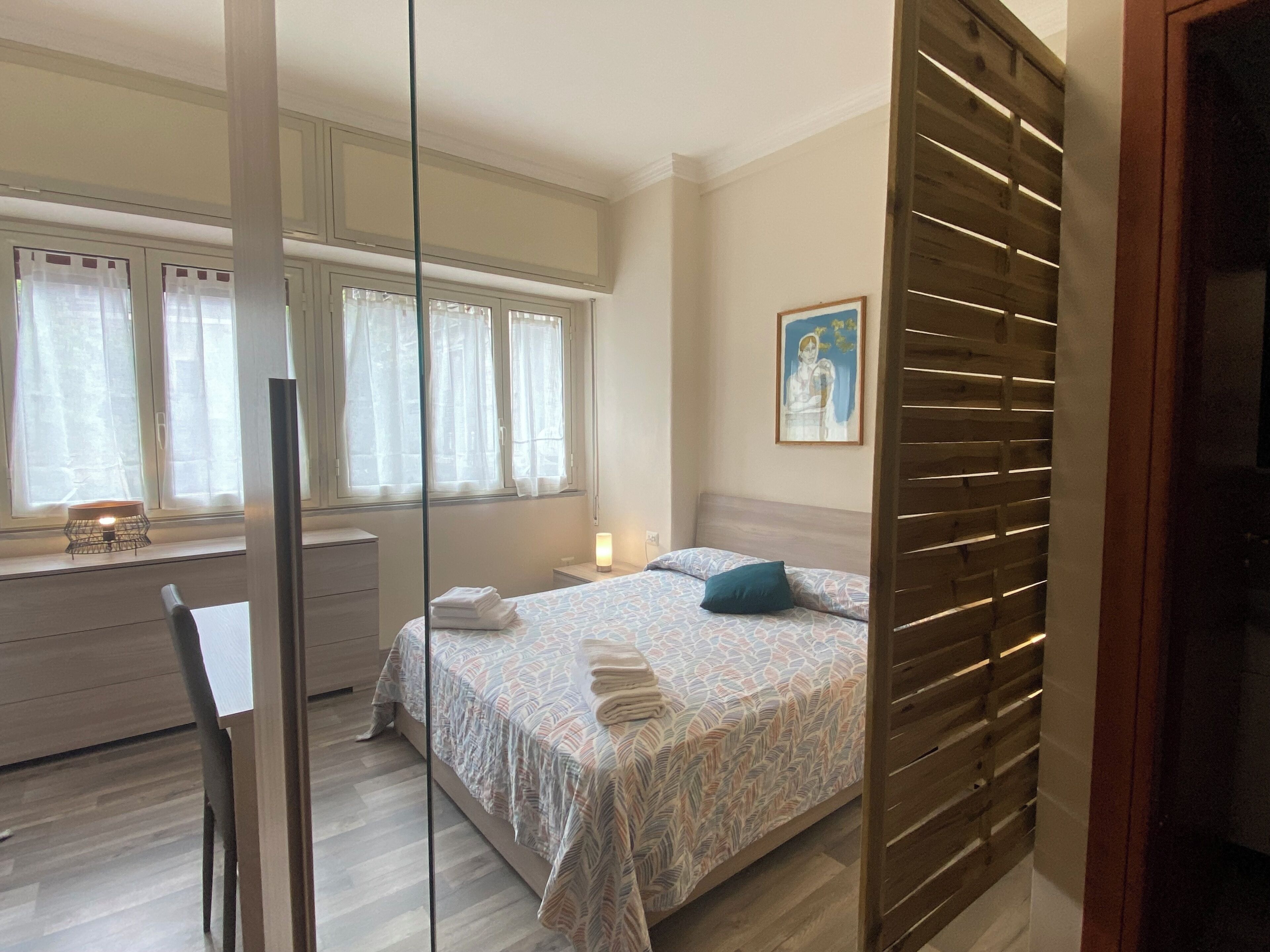 A bedroom with a bed, a desk, a chair, a window with curtains, a bedside table with a lamp, and a painting on the wall.