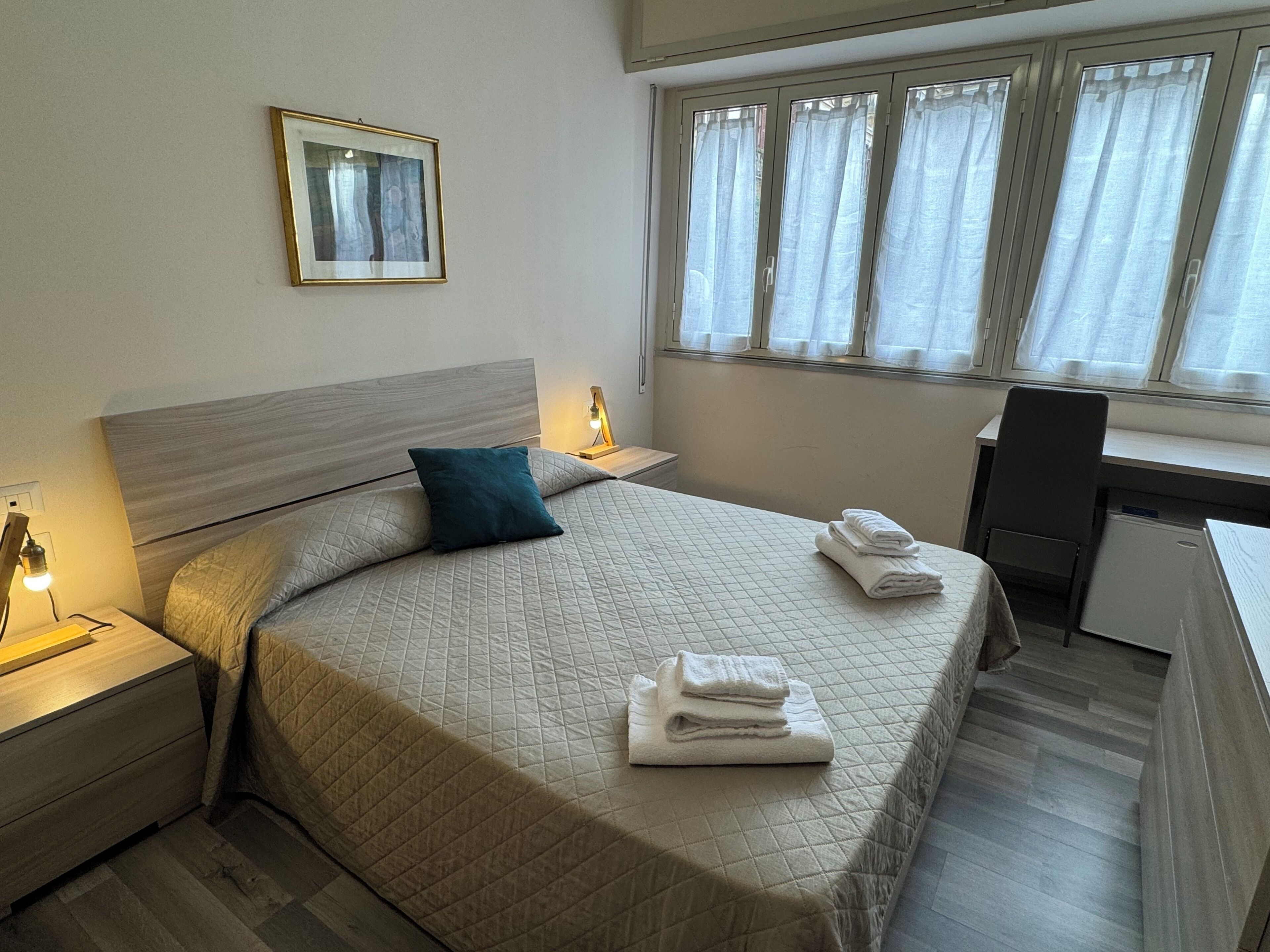 Een hotelkamer met een bed, een bureau, een stoel, een nachtkastje met een lamp, een raam met gordijnen en een lijst met een afbeelding aan de muur.
