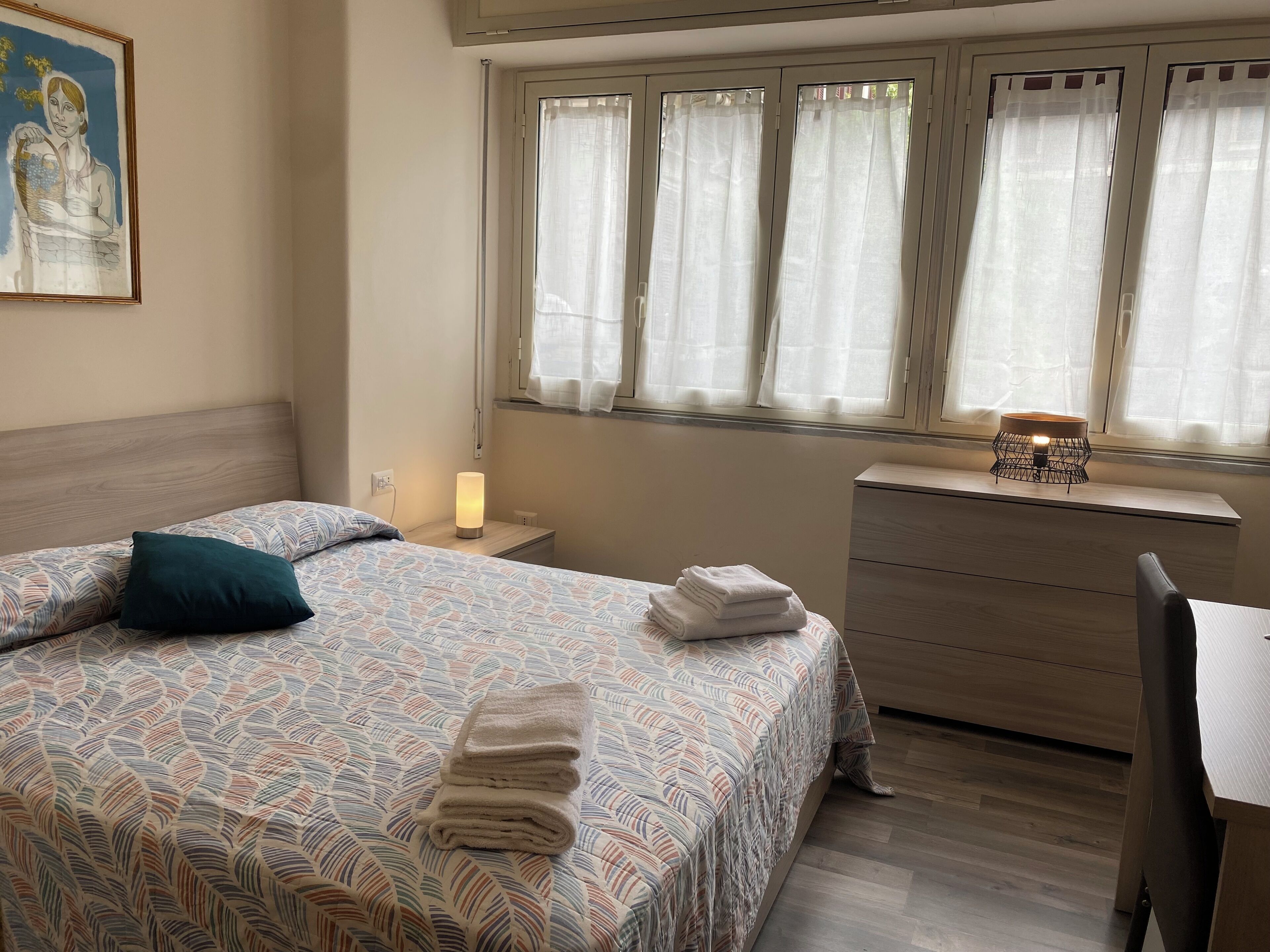 A bedroom with a bed, a nightstand, a lamp, a dresser, and a window with curtains.