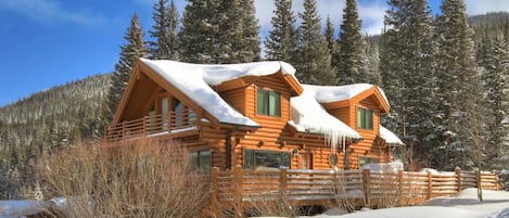 Una cabaña de troncos con techo cubierto de nieve, rodeada por una valla de madera y altos árboles de hoja perenne.