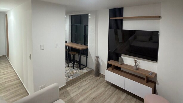 Uma sala moderna com um sofá, uma TV fixada na parede, um espaço de jantar com mesa e cadeiras, e um corredor que dá para outro quarto.