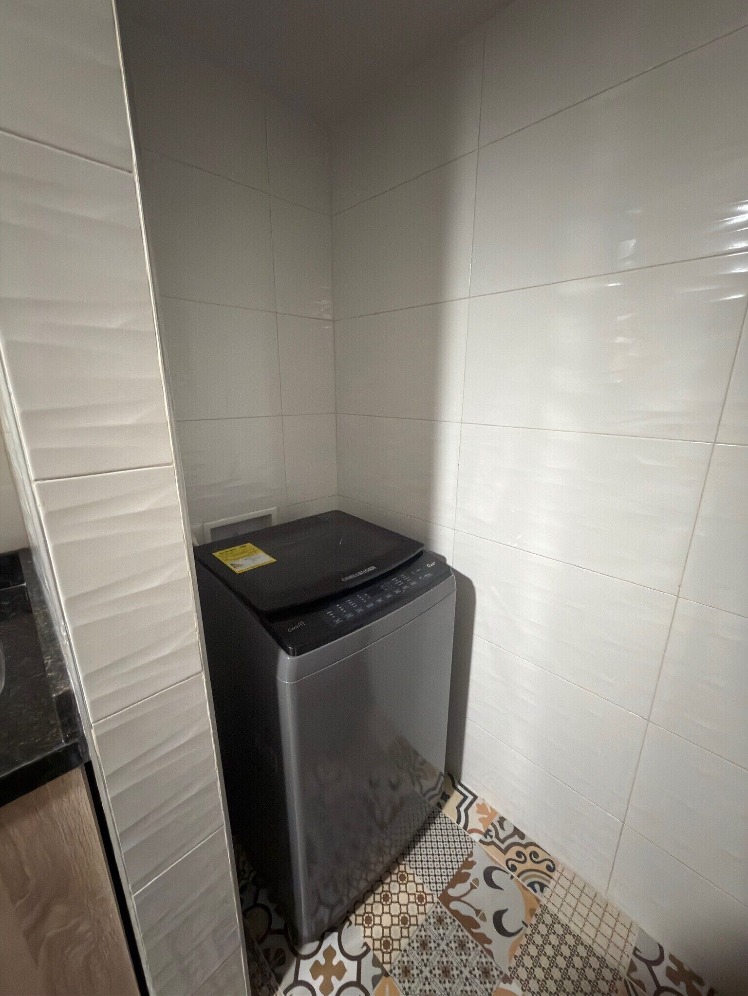 A compact, dark-colored washing machine in a corner of a room with white tiled walls and a patterned floor.