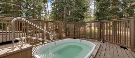 Un jacuzzi en una terraza de madera rodeado por una valla de madera, con un bosque al fondo.