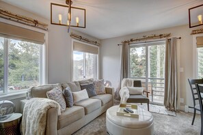 Ein geräumiges Wohnzimmer mit einem beigefarbenen Sofa, einem runden Hocker und einem Sessel. Große Fenster mit Vorhängen, eine Chandelierleuchtre und ein Spiegel an der Wand sind zu sehen.