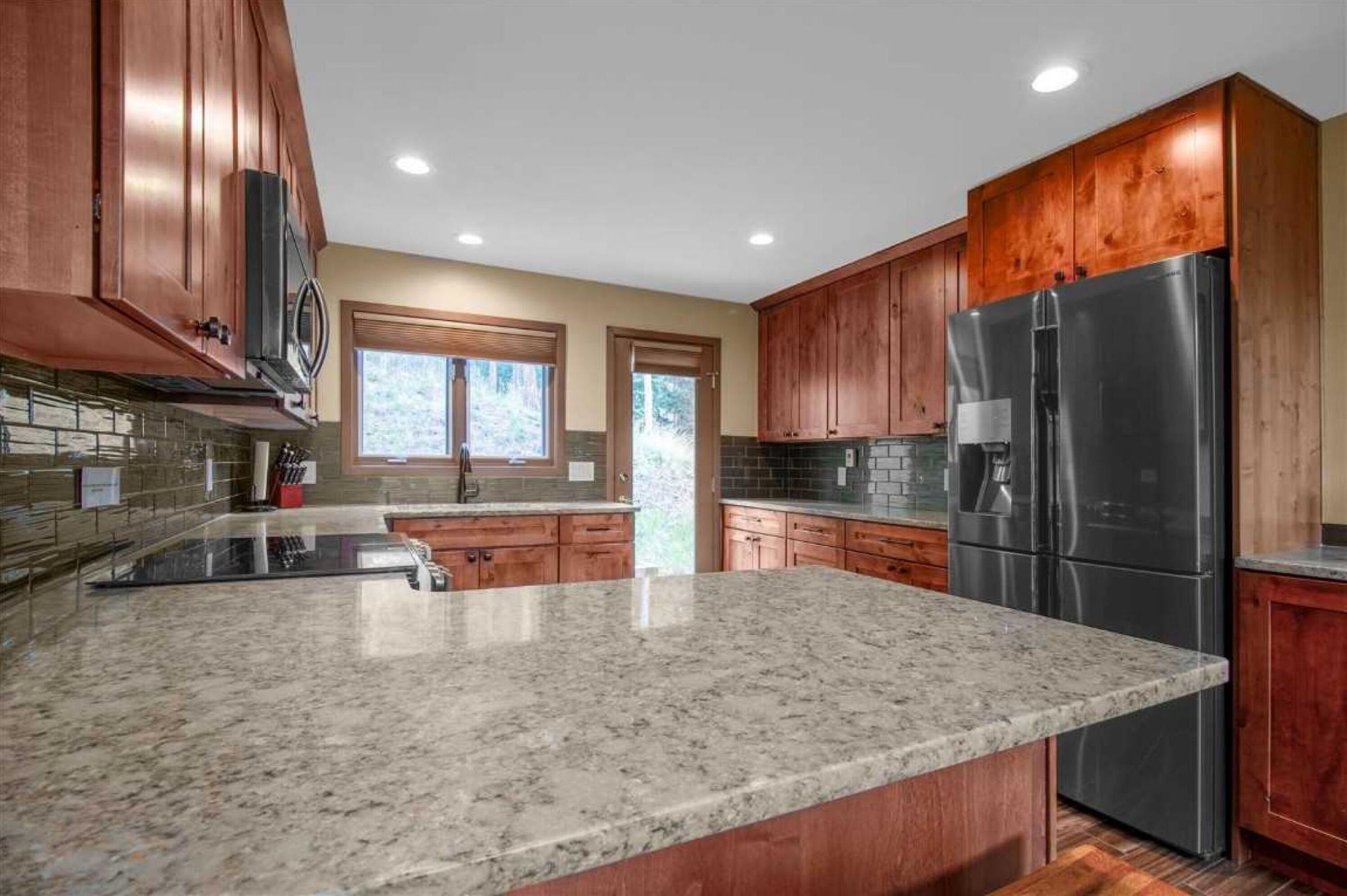 A kitchen with granite countertops, wooden cabinets, a stainless steel refrigerator, and a window with a view of trees.
