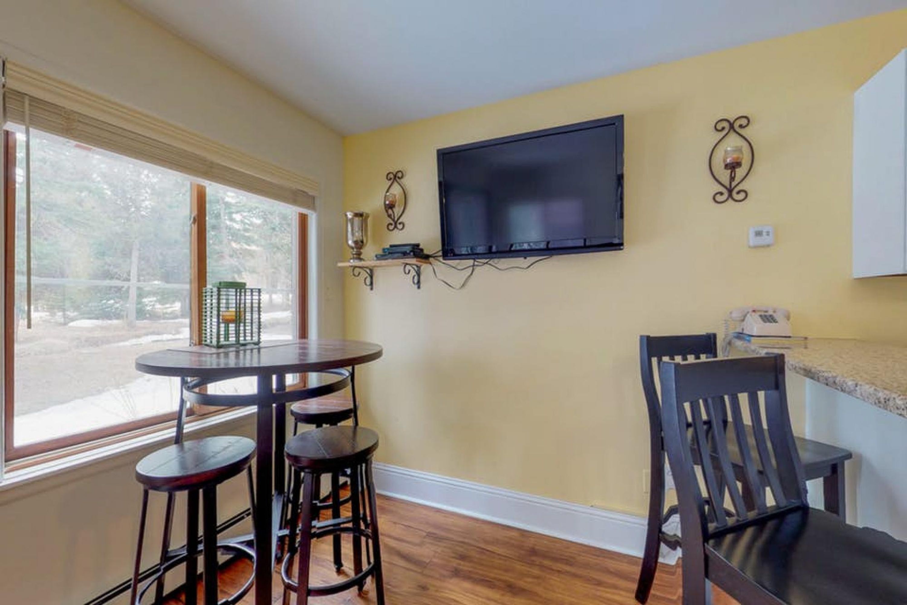 A dining area with a round table, two barstools, a television mounted on the wall, and a window with a view of trees.