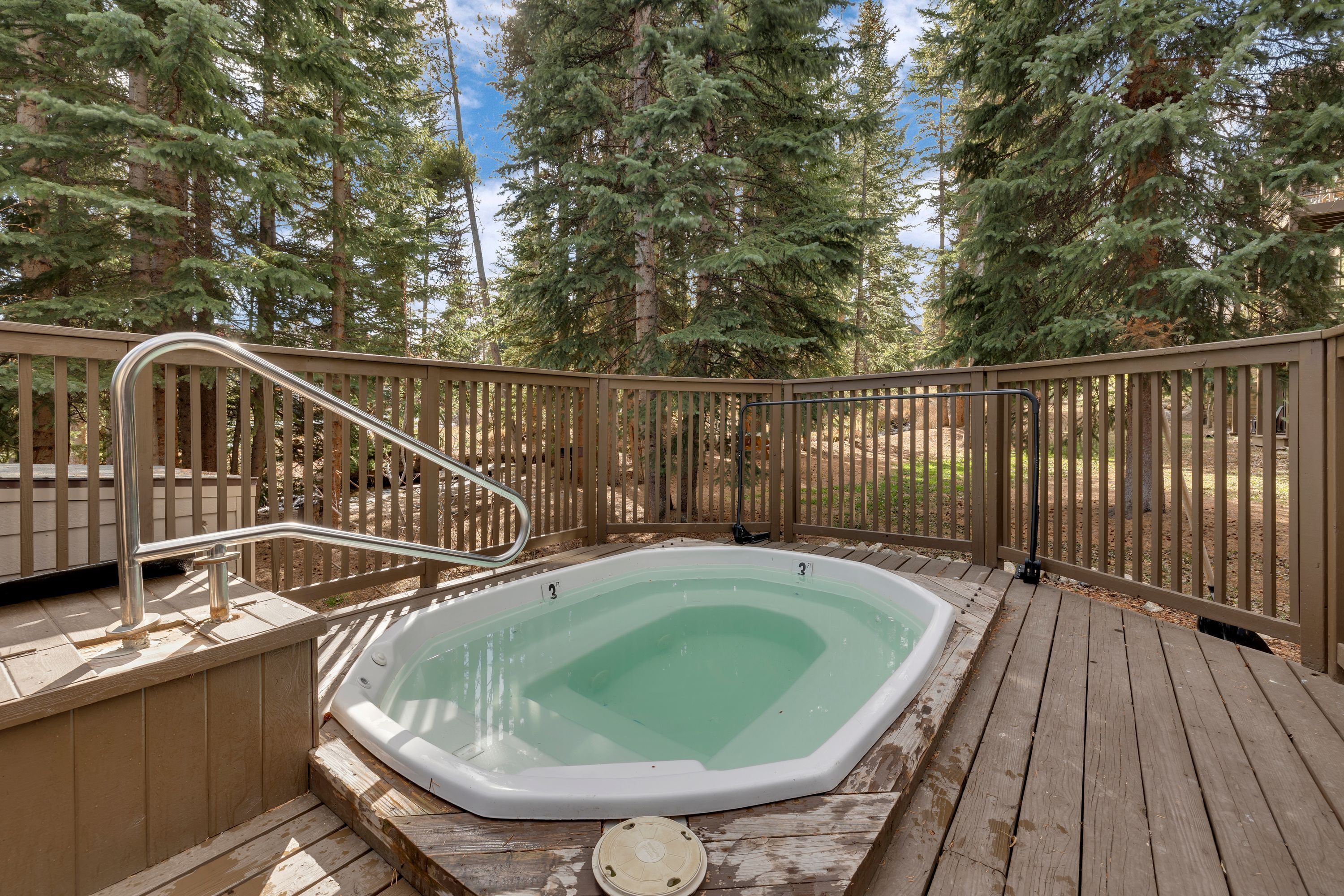 Ein Whirlpool auf einer Holzterrasse, umgeben von einem Holzzaun, mit einem Wald im Hintergrund.