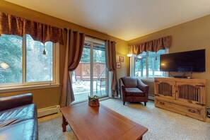 A living room with a sofa, a wooden coffee table, a television on a wooden stand, and large windows with curtains. - Wildwood Suites 101 in Breckenridge (Breckenridge)