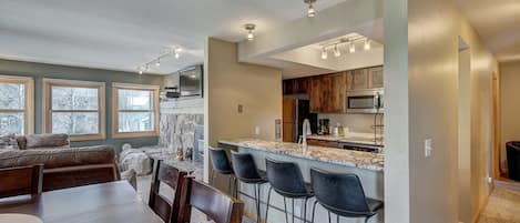 A modern kitchen with a bar area, a dining table with chairs, and a living area with a sofa and windows.