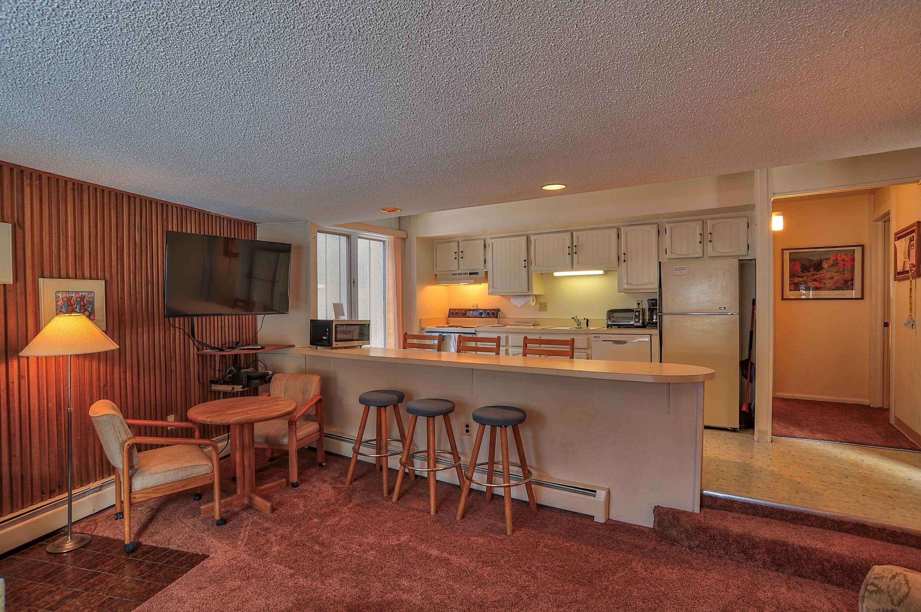 A compact kitchen with a central island, bar stools, a dining table, and a TV.