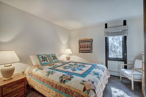 A bedroom with a bed, a nightstand, a lamp, a chair, a window, and a wall hanging. - Gold Camp II E87 in Breckenridge (Breckenridge)