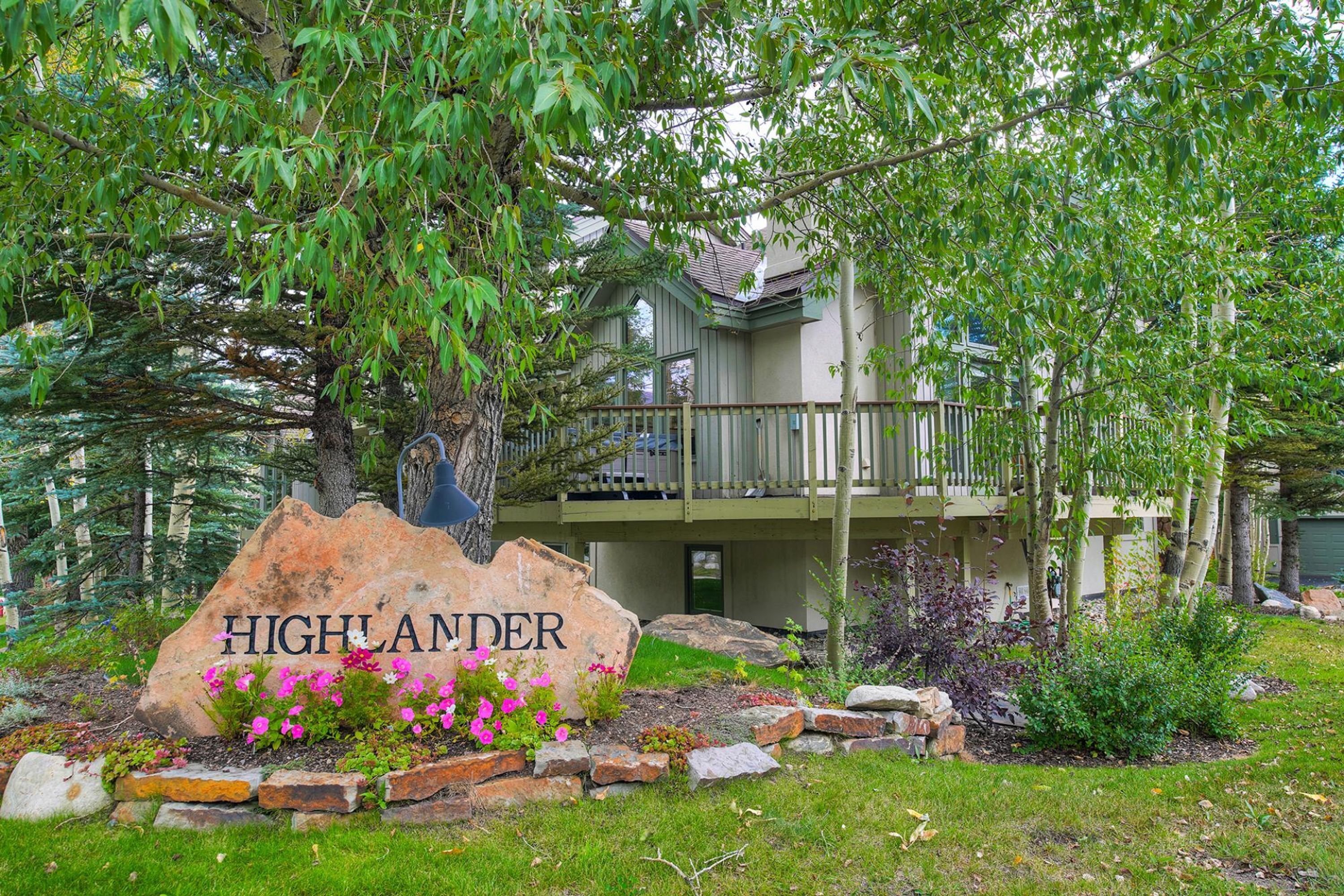A house with a stone sign labeled 'HIGHLANDER,' surrounded by a garden with pink flowers and a wooden deck.