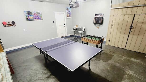 Une table de ping-pong se trouve dans une pièce dont le sol est en béton, où l’on aperçoit également une table de baby-foot et deux tableaux accrochés au mur.