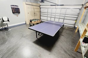 Ein Tischtennistisch in einer Garage mit Holztisch und Stühlen, einer Garagentür und einem an der Wand befestigten Gegenstand.