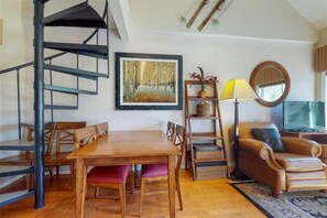 Una habitación con una mesa de comedor de madera, sillas, un sofá, una estantería, una lámpara, un espejo y una escalera de caracol.