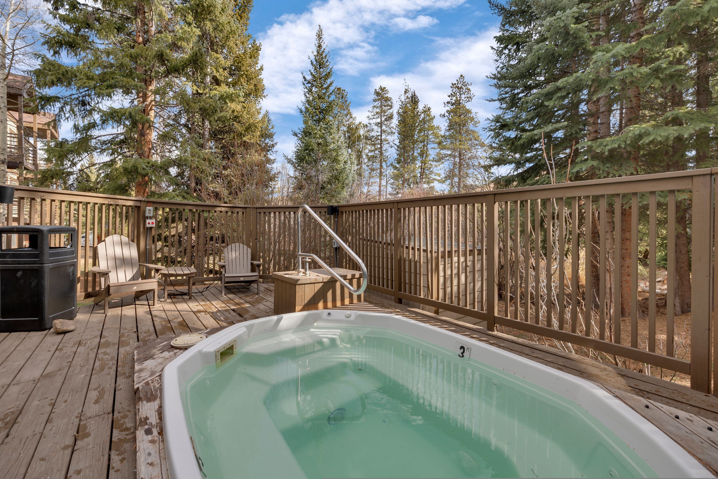 Een hottub omringd door een houten terras, met een houten hek en hoge bomen op de achtergrond.