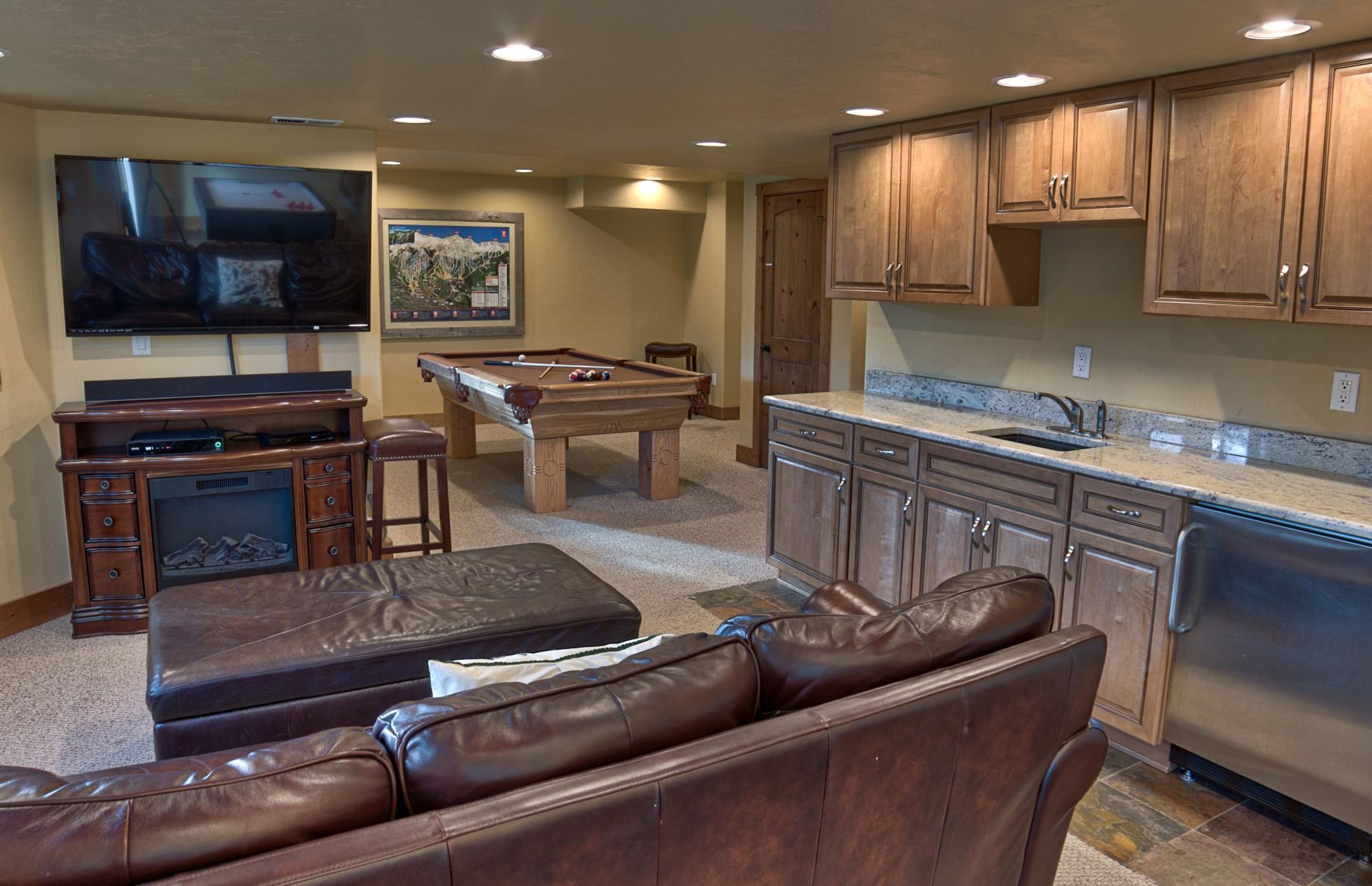 A living room with a flat-screen TV, a pool table, leather sofas, and a kitchen with wooden cabinets and a stainless steel dishwasher.