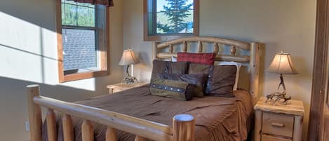 Una cama de madera con cabecera, dos mesitas de noche con lámparas, una ventana con vistas a árboles y una cortina.