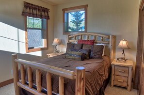 A wooden bed with a headboard, two bedside tables with lamps, a window with a view of trees, and a curtain. - 120 Rachel Lane in Breckenridge (Breckenridge)