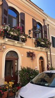 Un edificio con facciata marrone, numerosi balconi fioriti e un'auto bianca parcheggiata davanti.