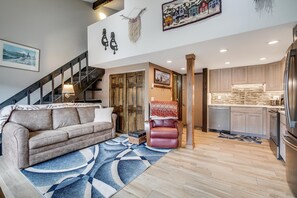 Ein modernes Wohnzimmer mit einer Couch, einem Teppich mit Muster, einer Treppe und einem Küchenbereich mit Holzschränken und einem Kühlschrank.