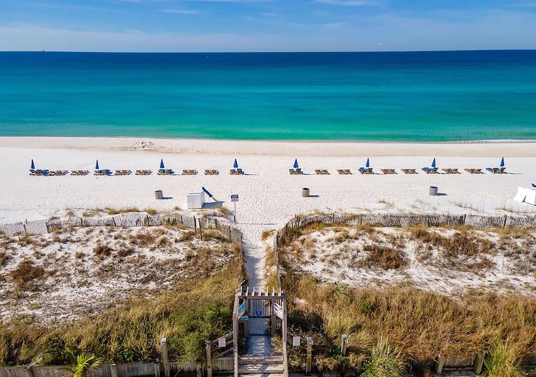 Een strand met rijen ligbedden en parasols, een houten looppad naar het strand, en een helder uitzicht op de oceaan.