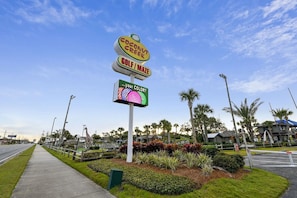 Ein Schild mit 'Coconut Creek Golf/Mazda' und einer digitalen Anzeige mit der Aufschrift 'deine FARBE?', im Hintergrund Palmen und eine Giraffe.