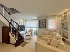 A spacious living room with a staircase, a white sofa set, a bookshelf, and a large window with curtains. - Tutto Bene Apartment 6906 (Helgueras)