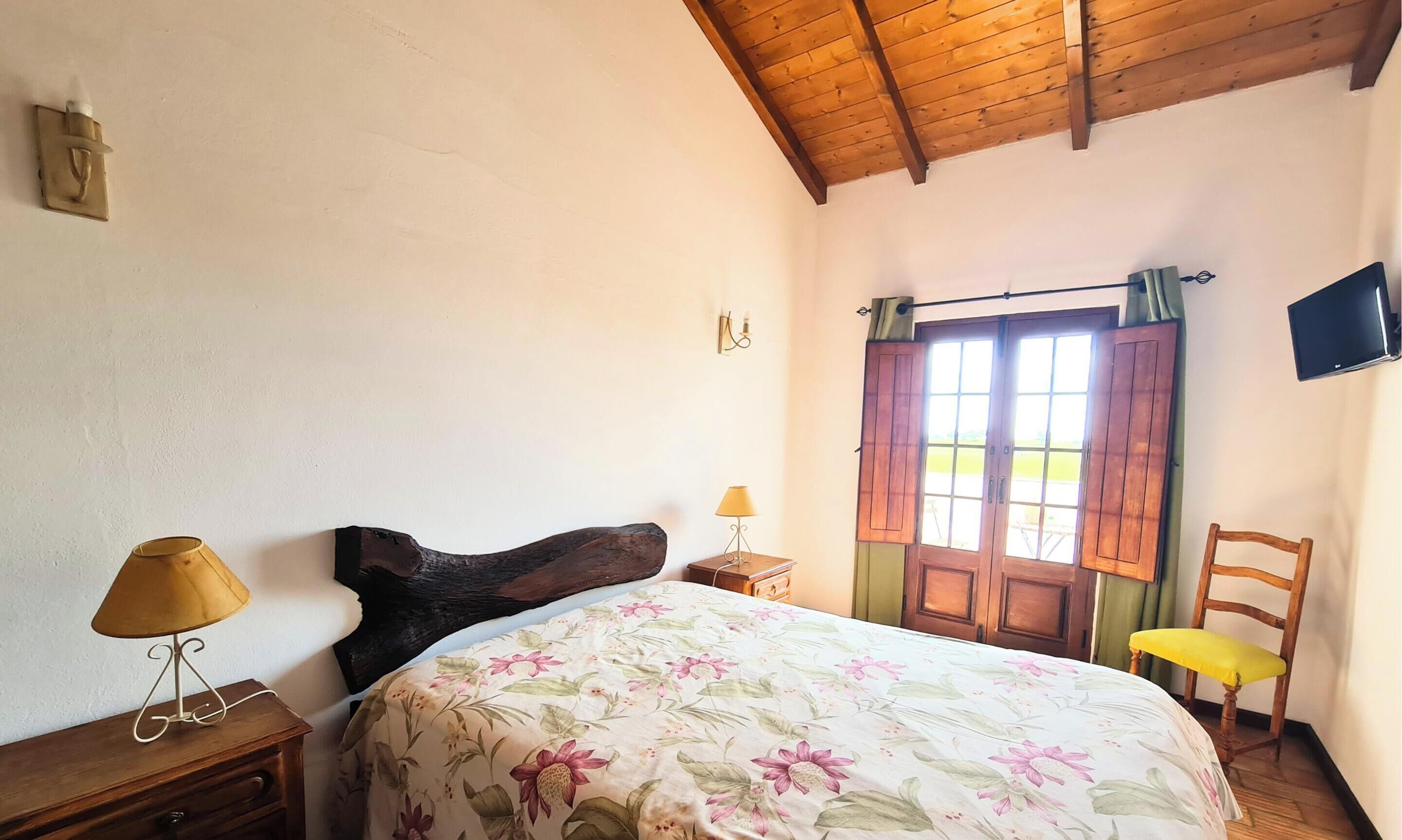 Une chambre à coucher avec un lit, une tête de lit en bois, une table de chevet avec une lampe, une chaise et un téléviseur fixé au mur.