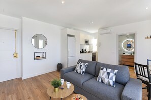 Een moderne woonkamer met een grijze bank, twee decoratieve kussens, een kleine ronde salontafel met een plant en twee kopjes, en een houten vloer.