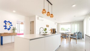Moderni keittiö, jossa on keskeinen saareke, riippuvalaisimet sekä ruokailutila tuoleineen ja pöytineen.