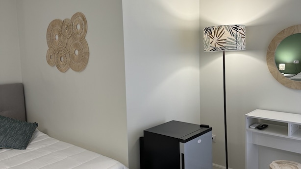 Ein Schlafzimmer mit einem Bett, einem kleinen schwarzen Kühlschrank, einer Stehlampe, einem weißen Schreibtisch und einem runden Spiegel.